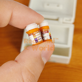 Two miniature orange prescription pill bottles with detailed printed labels held between fingers on a wooden surface, caps removed and placed beside them, miniature first aid box visible in background