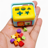 Miniature working shape sorter with yellow lid and blue body resting on an open palm, with 10 tiny colorful shape pieces — stars, cubes, cylinders, triangles — scattered in the hand below it
