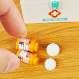 Two miniature orange prescription pill bottles with detailed printed labels held between fingers on a wooden surface, caps removed and placed beside them, miniature first aid box visible in background