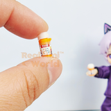 Two miniature orange prescription pill bottles with detailed printed labels held between fingers on a wooden surface, caps removed and placed beside them, miniature first aid box visible in background