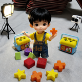 Miniature working shape sorter with yellow lid and blue body resting on an open palm, with 10 tiny colorful shape pieces — stars, cubes, cylinders, triangles — scattered in the hand below it