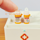 Two miniature orange prescription pill bottles with detailed printed labels held between fingers on a wooden surface, caps removed and placed beside them, miniature first aid box visible in background