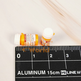 Two miniature orange prescription pill bottles with detailed printed labels held between fingers on a wooden surface, caps removed and placed beside them, miniature first aid box visible in background