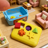 Miniature working shape sorter with yellow lid and blue body resting on an open palm, with 10 tiny colorful shape pieces — stars, cubes, cylinders, triangles — scattered in the hand below it
