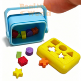 Miniature working shape sorter with yellow lid and blue body resting on an open palm, with 10 tiny colorful shape pieces — stars, cubes, cylinders, triangles — scattered in the hand below it